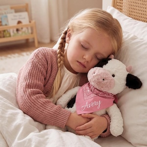 May include: A plush cow stuffed animal with white and black fur, featuring a pink snout and small pink ears. The cow wears a pink bandana with a white paisley pattern and the name Avery embroidered in white script across the fabric.