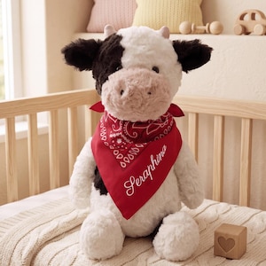 May include: A plush cow stuffed animal with black and white fur, featuring a light pink muzzle and small horns. The cow wears a red bandana with a white paisley pattern and the name Seraphina embroidered in white script across the fabric.