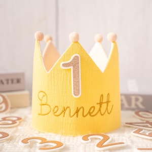 Può includere: Una corona di feltro giallo con il numero "1" in glitter dorati e il nome "Bennett" ricamato in oro. La corona ha sei punte, ognuna sormontata da un piccolo pompon chiaro. Progettata per una festa di primo compleanno.