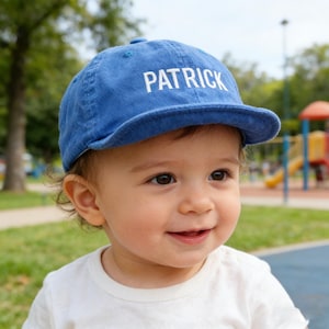 Könnte beinhalten: Blaue Baseballkappe mit dem Namen "PATRICK" in weißen Blockbuchstaben. Die Kappe wird von einem lächelnden Kind in einem weißen T-Shirt getragen, mit einem Parkhintergrund.