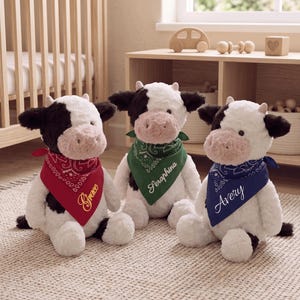 May include: Three plush cow toys with white and black fur, pink muzzles, and small horns. Each cow wears a bandana: red with the name Grace, green with Seraphina, and blue with Avery. The bandanas feature a white paisley pattern.