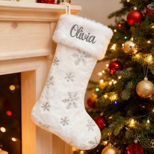 May include: A white Christmas stocking with a fluffy cuff and snowflake pattern. The name "Olivia" is embroidered in grey script on the cuff. The stocking is displayed near a decorated Christmas tree.
