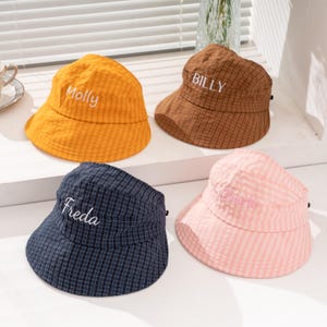 May include: Four bucket hats in yellow, brown, navy blue, and light pink, each featuring a subtle checkered pattern. The yellow hat is embroidered with the name Molly, the brown hat with Billy, the navy hat with Freda, and the pink hat with Charli.