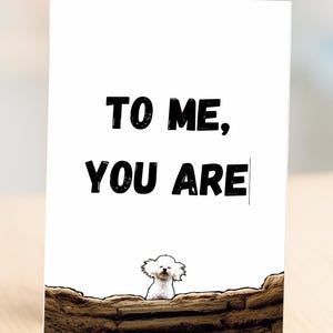 May include: A white greeting card with the text "TO ME, YOU ARE" in bold black letters. A small white dog peeks over a brown wall at the bottom of the card. The card is on a light wooden surface.