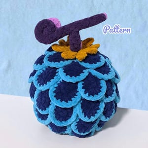 Puede incluir: Una fruta de ganchillo hecha a mano en tonos azules, con un tallo morado oscuro y una base amarilla. La fruta está cubierta de patrones superpuestos en forma de escamas. La palabra "Pattern" está escrita en morado claro.
