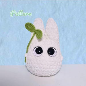 May include: A handmade white crochet plush toy with large black eyes and a green sprout. The word "Pattern" is written in a light green script in the upper left corner. The plush is set against a light blue background.