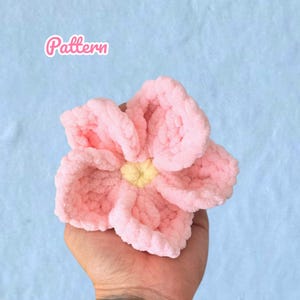 Könnte beinhalten: Eine handgefertigte, plüschige rosa Blume mit gelber Mitte. Die Blume wird in einer Hand vor einem hellblauen Hintergrund gehalten. Das Wort "Pattern" steht in rosa Schrift oben links.