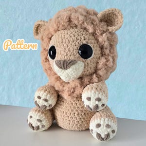 Könnte beinhalten: Ein handgefertigtes gehäkeltes Löwen-Plüschtier in Beige- und Cremetönen. Der Löwe hat große schwarze Augen, eine flauschige Mähne und detaillierte Pfoten. Das Wort "Pattern" ist in Schreibschrift geschrieben.