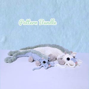 Könnte beinhalten: Zwei gehäkelte Drachen-Plüschtiere. Einer ist hellblau und der andere weiß. Beide haben graue Akzente und große schwarze Augen. Der Text "Pattern Bundle" befindet sich oben in der Mitte des Bildes.