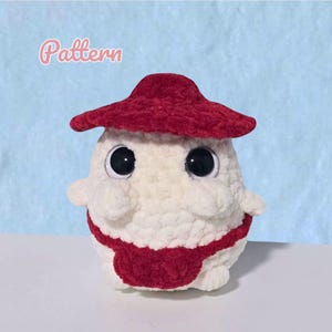 Könnte beinhalten: Ein handgefertigtes, pilzförmiges Kuscheltier. Es ist hauptsächlich weiß mit einem roten Hut und einem roten Band. Es hat große Augen mit schwarzen Pupillen und kleine Arme. Das Wort "Pattern" steht in rosa Schrift oben links.