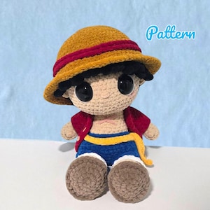 Op de afbeelding: Gehaakte pop van een stripfiguur met een gele hoed met een rode band, een rood jasje, een blauwe korte broek met een gele streep en bruine schoenen. De pop heeft grote zwarte ogen en zit. Het woord "Pattern" is in het blauw geschreven.