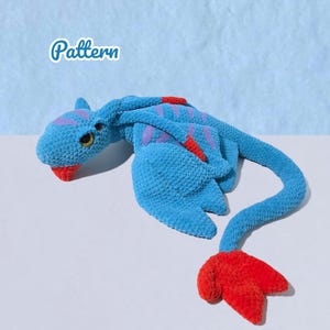 Könnte beinhalten: Ein handgefertigtes blaues Häkeldrachen-Spielzeug mit roten und lila Akzenten. Der Drache hat einen langen Schwanz und Flügel. Das Wort "Pattern" befindet sich in der oberen linken Ecke.
