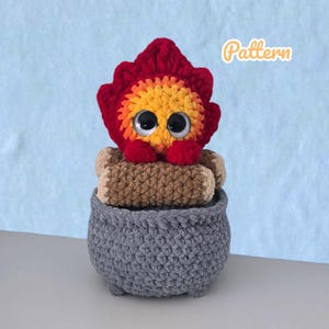 May include: A handmade crochet fire-themed toy. The toy features a red and yellow flame design with large googly eyes, sitting atop a stack of brown logs inside a gray crochet pot. The word "Pattern" is written in the upper right corner.