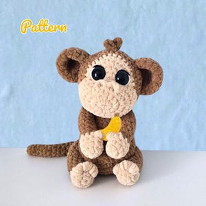 Könnte beinhalten: Eine gehäkelte Affenpuppe in Braun- und Beigetönen, die eine gelbe Banane hält. Der Affe hat große schwarze Augen und einen lächelnden Ausdruck. Das Wort "Pattern" ist in gelben Schreibschriftbuchstaben geschrieben.