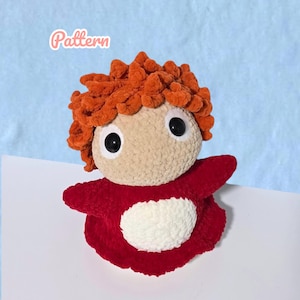 Könnte beinhalten: Eine handgefertigte Plüschpuppe mit hellbraunem Gesicht, großen schwarzen Augen und orangefarbenem, strukturiertem Haar. Die Puppe trägt ein rotes Outfit mit weißer Mitte. Das Wort "Pattern" steht in rosa Schrift oben links. Der Hintergrund ist hellblau und weiß.