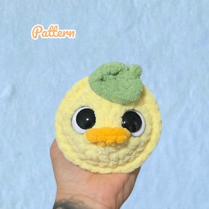 Könnte beinhalten: Eine handgefertigte gelbe Häkelente mit großen schwarzen Augen und einem orangefarbenen Schnabel. Ein grünes Blatt ist oben auf dem Kopf der Ente befestigt. Das Wort "Pattern" ist sichtbar.