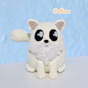 Peut inclure: Une peluche en forme de renard blanc crocheté, avec de grands yeux noirs et une poitrine blanche duveteuse. Le jouet a un petit nez noir et des oreilles intérieures de couleur beige. Le mot "Pattern" est écrit en orange en haut à droite.