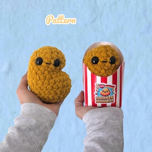 Könnte beinhalten: Zwei goldbraune, texturierte Häkel-Plüschtiere in Form von Chicken Nuggets mit schwarzen Sicherheitsaugen und kleinen gestickten Mündern. Ein Nugget wird einzeln gehalten, während das andere in einem rot-weiß gestreiften Pappkarton mit der Aufschrift I LOVE CHICKEN NUGGETS steckt.