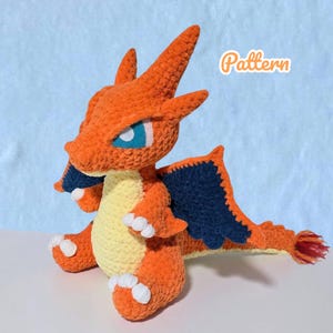 May include: Crocheted orange dragon plushie with blue wings and accents, and a yellow belly. The dragon has white claws and blue eyes. The word "Pattern" is written in the upper right corner.