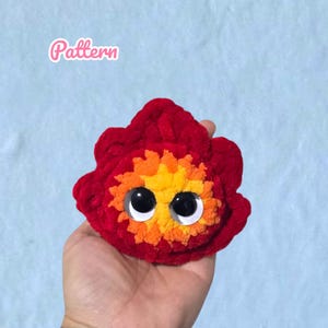 Könnte beinhalten: Ein handgefertigtes, plüschiges, rotes Geschöpf mit großen, schwarzen Wackelaugen und einem gelb-orangenen Zentrum. Das Wort "Pattern" ist in rosa Schreibschrift oben links geschrieben. Das Plüschtier wird in einer Hand vor einem hellblauen Hintergrund gehalten.