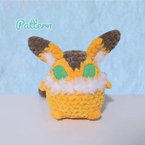 May include: A handmade, plush creature with a yellow body, brown and yellow ears, and green eyes. The creature has a white fluffy collar and is labeled with the word "Pattern" in a cursive font.