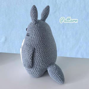 Może przedstawiać: Szara, szydełkowa pluszowa zabawka w kształcie postaci z kresk&oacute;wki z dużymi uszami, ogonem i białym brzuchem. Słowo "Pattern" jest napisane kursywą w prawym g&oacute;rnym rogu. Zabawka znajduje się na białej powierzchni z jasnoniebieskim tłem.