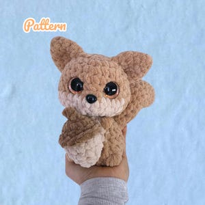 Könnte beinhalten: Ein gehäkeltes Eichhörnchen-Plüschtier in Brauntönen, das eine passende Eichel hält. Das Eichhörnchen hat große, glitzernde Augen und das Wort "Pattern" ist in orangefarbener Schrift geschrieben. Das Plüschtier wird vor einem hellblauen Hintergrund gehalten.