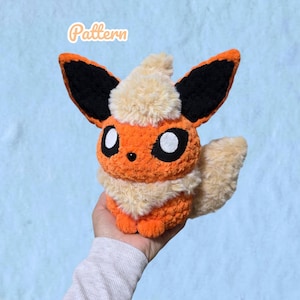 Könnte beinhalten: Ein Plüschtier, das einer fuchsartigen Kreatur aus orangefarbenem und cremefarbenem Garn ähnelt, mit großen spitzen Ohren mit schwarzen Innenteilen, einer flauschigen cremefarbenen Mähne und einem buschigen Schwanz. Das Wort Pattern steht in Weiß darüber.