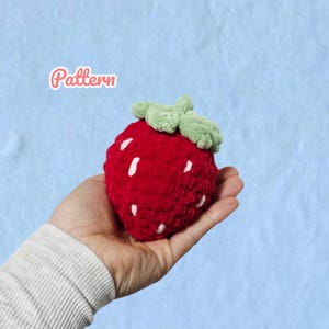 May include: A handmade, plush strawberry in vibrant red with white seed details and a green leafy top. The word "Pattern" is written in a pink cursive font. The strawberry is held in a hand against a light blue background.