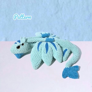 Könnte beinhalten: Ein gehäkelter Drache in Hellblau- und Dunkelblautönen. Der Drache hat einen langen Schwanz, Flügel und einen detaillierten Kopf mit kleinen Augen. Das Wort "Pattern" steht kursiv in der oberen linken Ecke.
