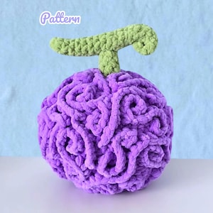 May include: A plush, round, purple fruit with a green stem. The fruit is made of a soft, textured material, and the word "Pattern" is written in a cursive font in the upper left corner.