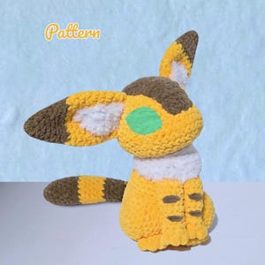 May include: A handmade crochet plushie in yellow, brown, and white. The plushie has large ears, a tail, and a green eye. The word "Pattern" is written in yellow script in the upper left corner. The plushie is sitting on a white surface with a light blue background.