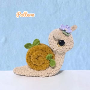 May include: A crocheted snail toy in beige, with a brown shell and green leaves. The snail has large, expressive eyes and a small purple flower on its head. The word "Pattern" is written in orange script.