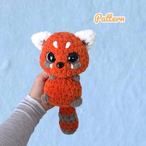 Könnte beinhalten: Ein handgefertigtes, gehäkeltes Rotpanda-Plüschtier in Orange und Weiß mit grauem Schwanz und Akzenten. Das Kuscheltier hat große, glitzernde Augen und wird in einer Hand gehalten. Das Wort "Pattern" steht in orangefarbener Schrift. Ca. 15 cm groß.