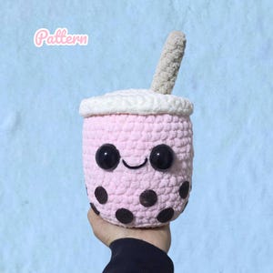 Könnte beinhalten: Ein gehäkeltes Plüschtier in Form eines Bubble-Tea-Bechers mit hellrosa Körper, weißem Deckel und beigem Strohhalm. Der Becher hat große schwarze Sicherheitsaugen aus Kunststoff, ein aufgesticktes schwarzes Lächeln und mehrere schwarze, kreisförmige Filzpunkte. Das Wort Pattern ist in der oberen linken Ecke zu sehen.