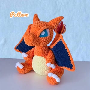 May include: A handmade orange crochet Charizard plushie with blue wings and white accents. The plushie has a yellow belly and a flame on its tail. The word "Pattern" is written in a cursive font in the upper left corner.