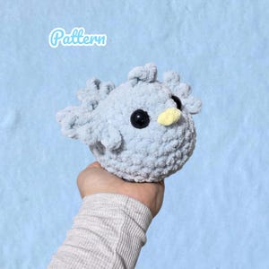 Könnte beinhalten: Ein hellblaues, gehäkeltes, vogelförmiges Plüschtier mit schwarzen Augen und einem gelben Schnabel. Das Wort "Pattern" ist in einer hellblauen Schrift geschrieben. Der Vogel hat kleine, abgerundete Flügel und ein strukturiertes, flauschiges Aussehen.
