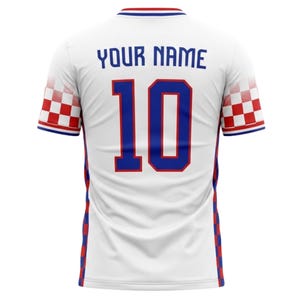 May include: White soccer jersey with blue and red accents. The jersey features the number 10 in blue, with the text "YOUR NAME" above it. The sleeves have a red and white checkered pattern.