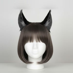 May include: A white mannequin head wearing a short, straight, brown bob wig with fringe. Attached to the top of the wig are two pointed, upright, black animal-style ears with grey inner detailing, creating a distinct feline or creature-like appearance.