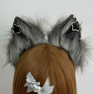 Puede incluir: Diadema con orejas de gato de piel sintética gris con detalles de estrellas y cadenas colgantes. Las orejas están unidas a una diadema negra. Un lazo plateado con alas está unido a la peluca. La peluca es marrón.