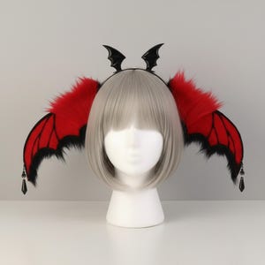 May include: A headband featuring two large red bat wings with black faux fur trim and small black dangling crystal-like pendants at the tips. The headband is adorned with two small black bat wing accents at the top center, worn on a white mannequin head.