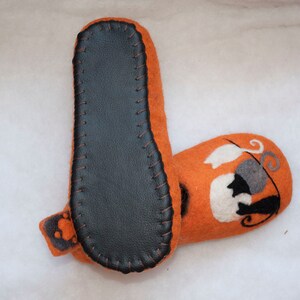 May include: Orange felt slippers with a black leather sole and a white and gray cat design on the top.