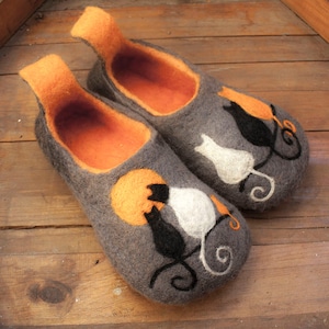May include: A pair of gray felt slippers with orange felt lining. The slippers feature a design of two cats, one black and one white, sitting on a branch with a yellow moon in the background.