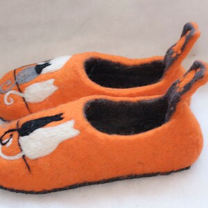May include: A pair of orange felt slippers with black and white cat designs. The slippers have a black felt lining and a black felt trim around the edges.