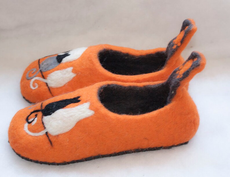May include: A pair of orange felt slippers with black and white cat designs. The slippers have a black felt lining and a black felt trim around the edges.
