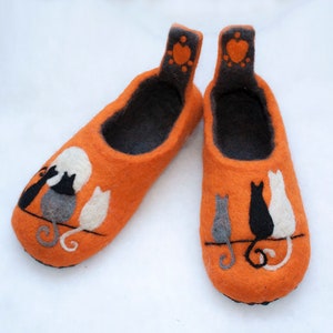 May include: A pair of orange felt slippers with a black, white, and gray cat design. The cats are sitting on a line with their tails curled. The slippers have a black felt lining and a small heart with paw prints on the back.