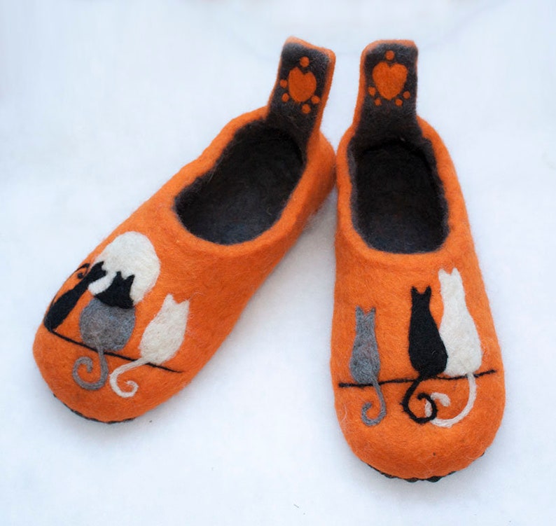 May include: A pair of orange felt slippers with a black, white, and gray cat design. The cats are sitting on a line with their tails curled. The slippers have a black felt lining and a small heart with paw prints on the back.
