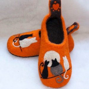 May include: A pair of orange felt slippers with black cat silhouettes and a white heart on the back. The slippers have a black felt lining.