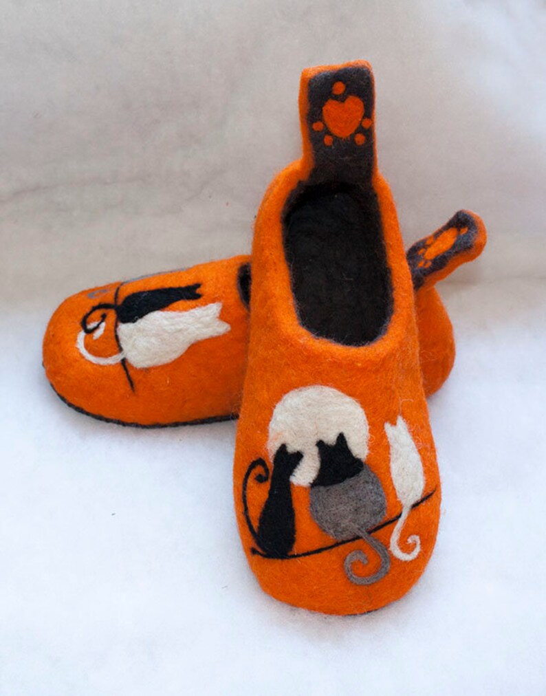 May include: A pair of orange felt slippers with black cat silhouettes and a white heart on the back. The slippers have a black felt lining.