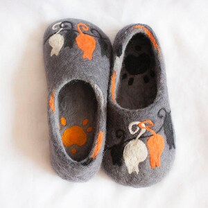 May include: A pair of gray felt slippers with orange, white, and black cat designs. The slippers have a paw print on the inside.